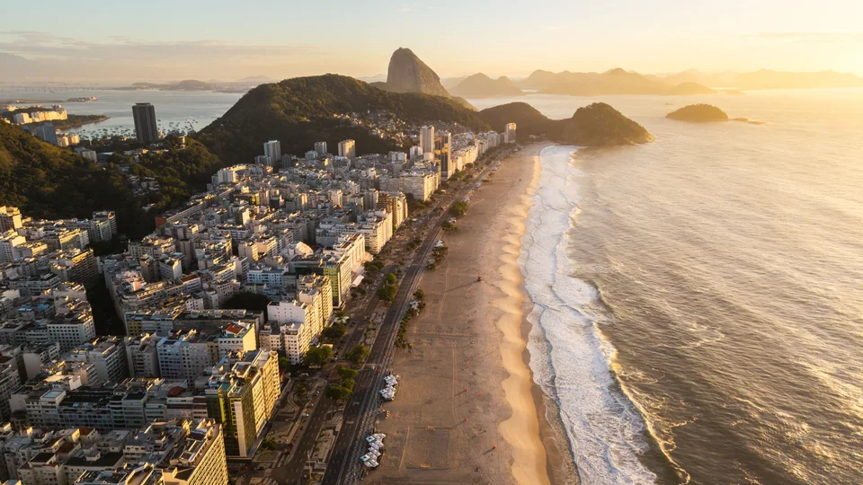 Aerial view along a long sandy beach with waves, rows of city buildings and mountains in warm evening light