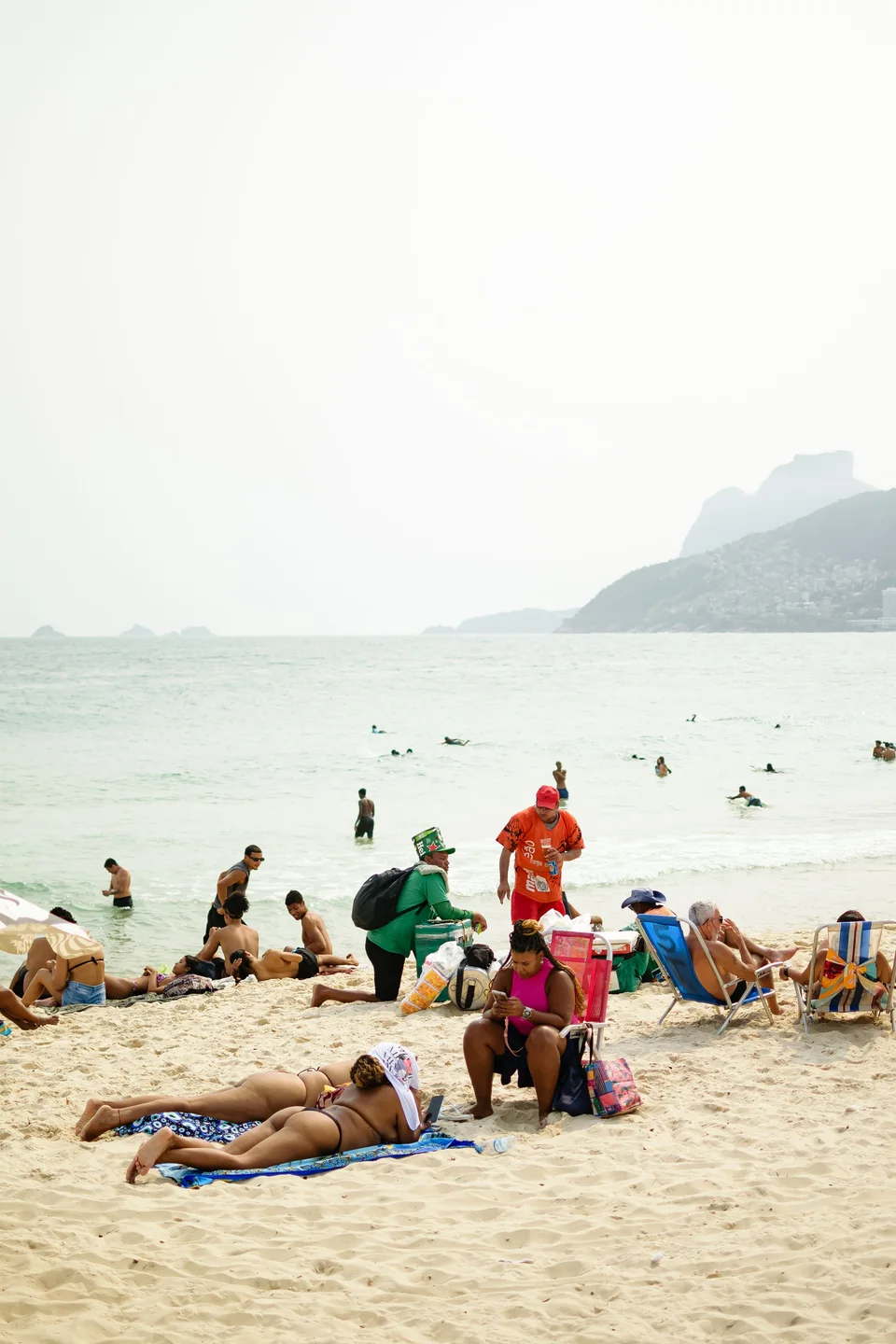 People resting on towels and beach chairs, vendors walking on the sand and swimmers in the sea beneath hazy mountains