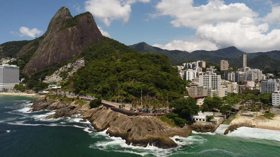 Green rocky mountains rising behind seaside buildings, a small sandy beach and waves along a rocky coast
