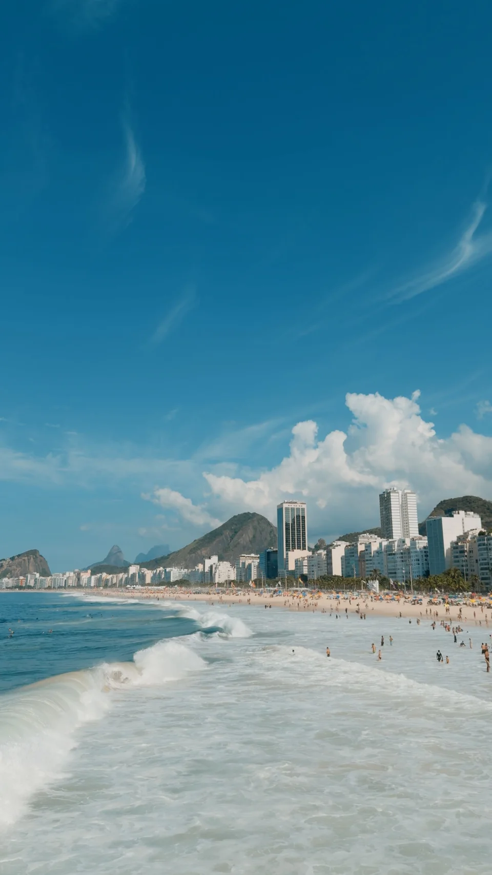 Foamy waves washing onto a broad sandy beach with people, beachfront towers and blue sky