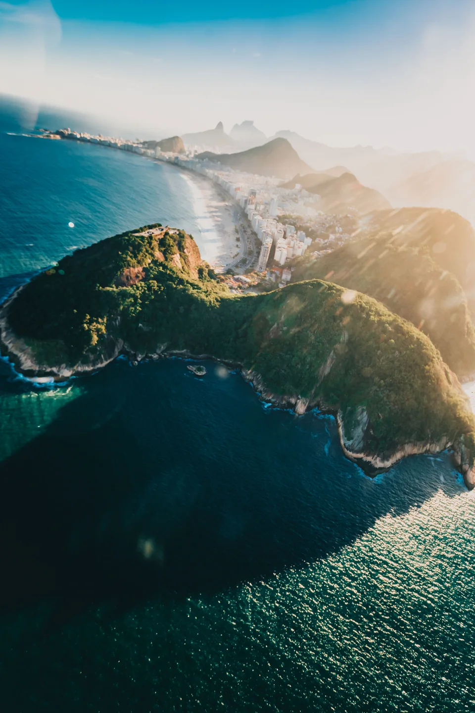 Aerial view of a steep green summit beside deep blue water and a long curving urban beach