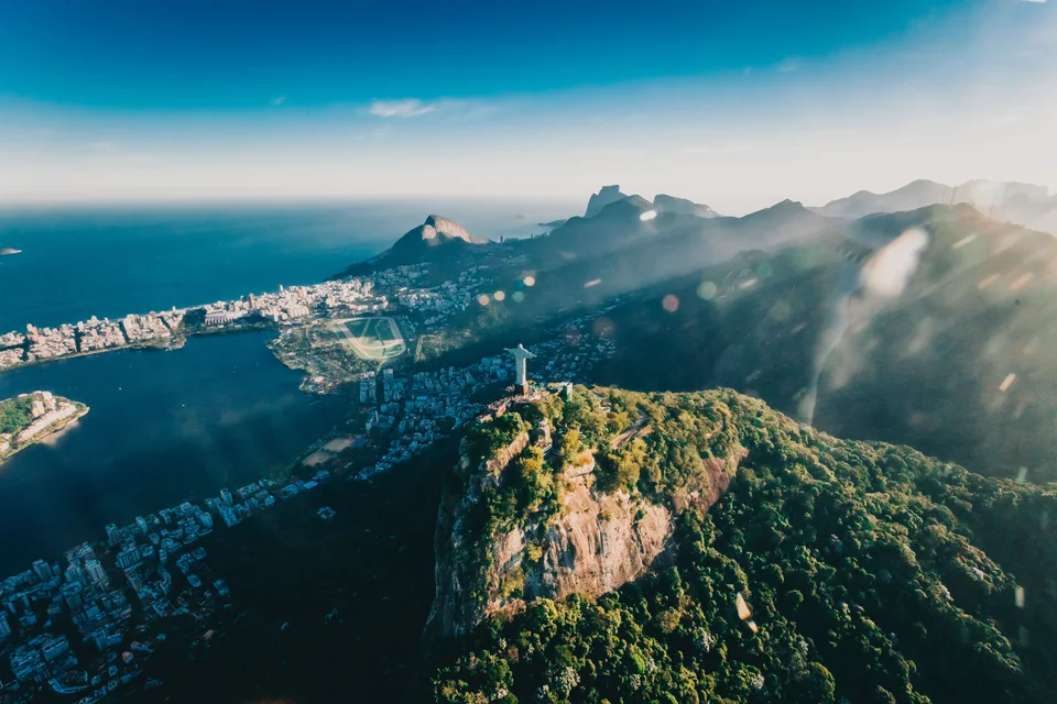Sunlit rocky green summit above a coastal city, blue ocean and mist drifting across nearby mountains