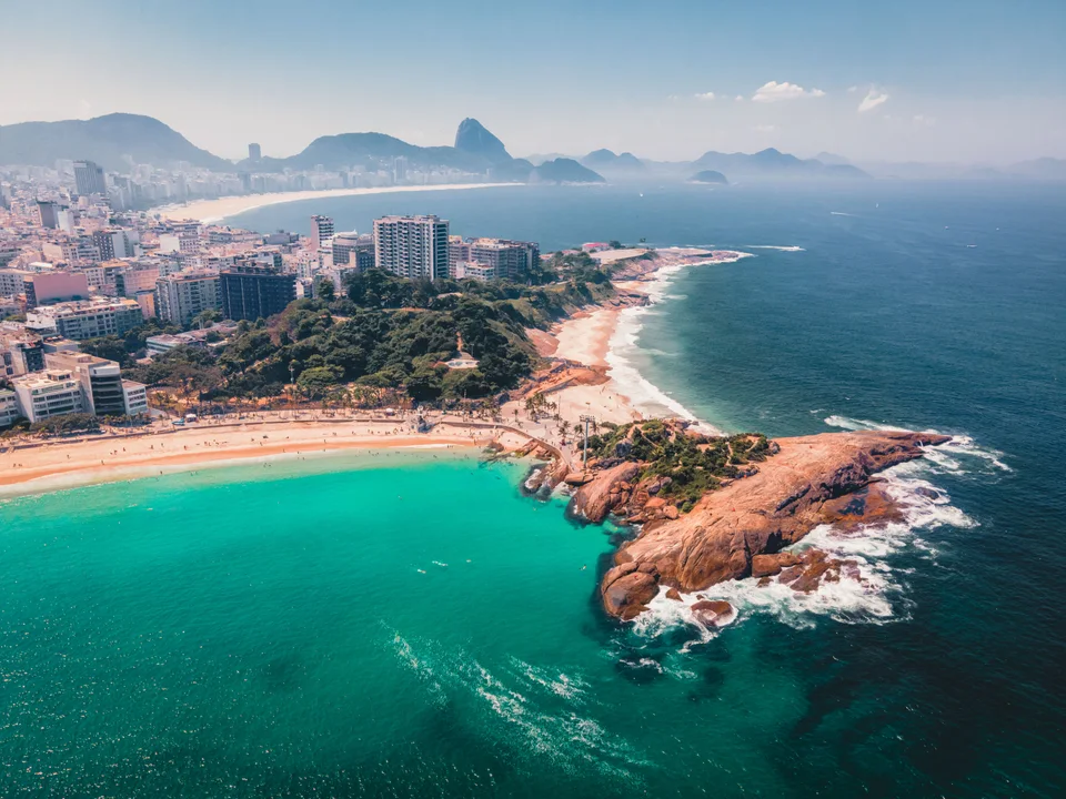 Aerial view of turquoise ocean beside a rocky headland, curving sandy beaches and rows of city buildings