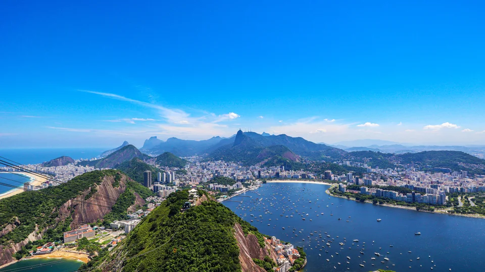 Panoramic view of a blue lagoon, green rocky hills, city buildings and mountains beneath bright blue sky