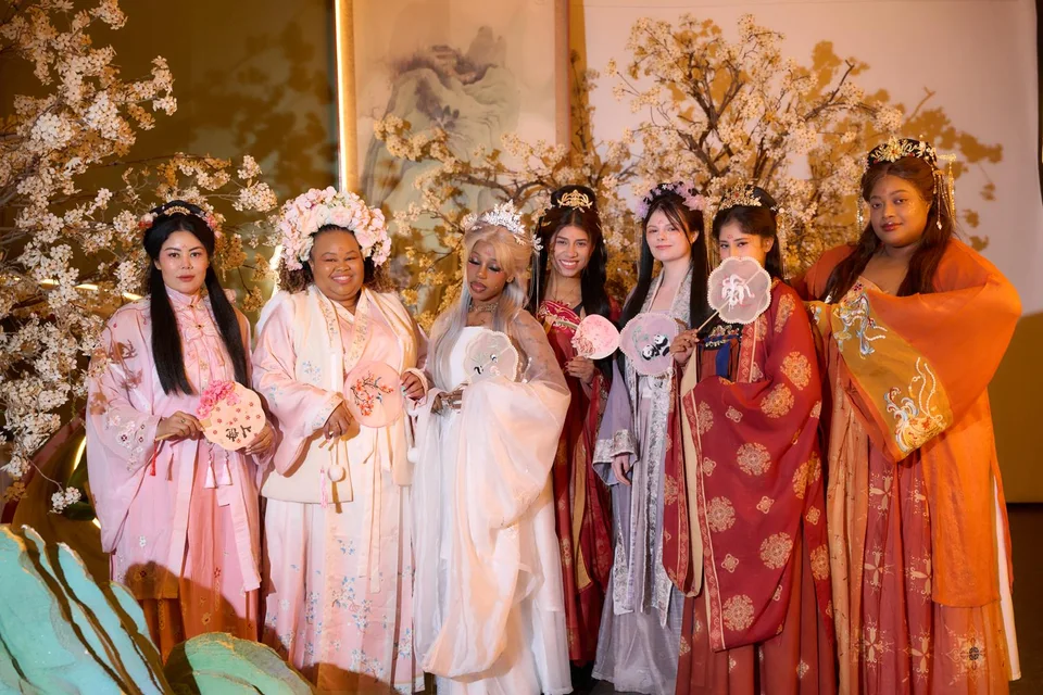 Seven travelers in colorful traditional Chinese dress, holding fans beside blossom decorations