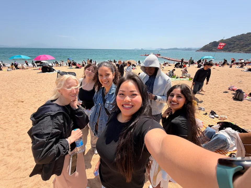 Friends taking a group selfie on a busy sandy beach beside turquoise water