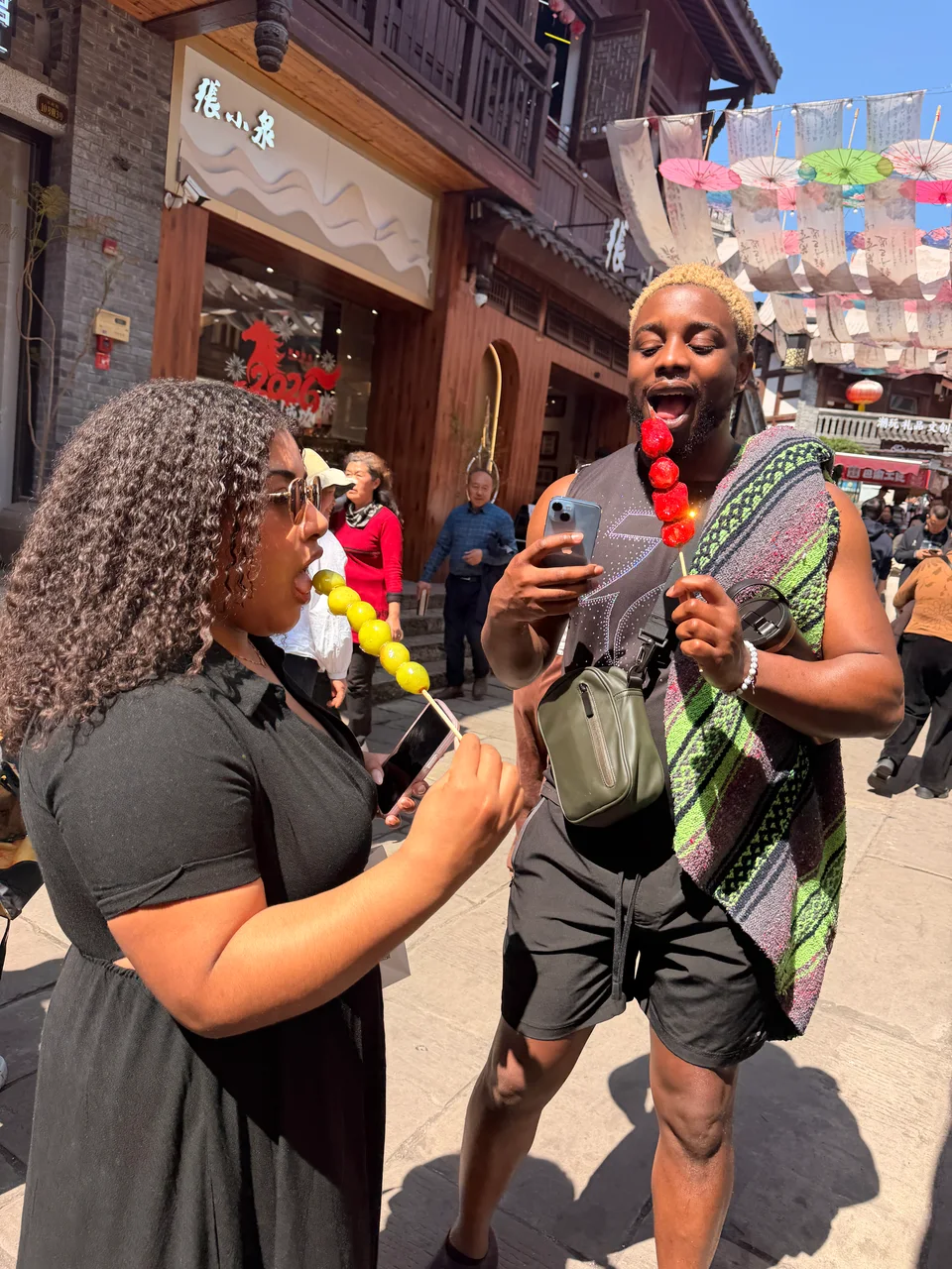 Two travelers enjoying skewered snacks on a lively street lined with wooden buildings