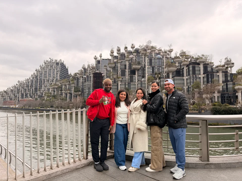 Five travelers posing beside a waterfront railing in front of a stepped concrete building