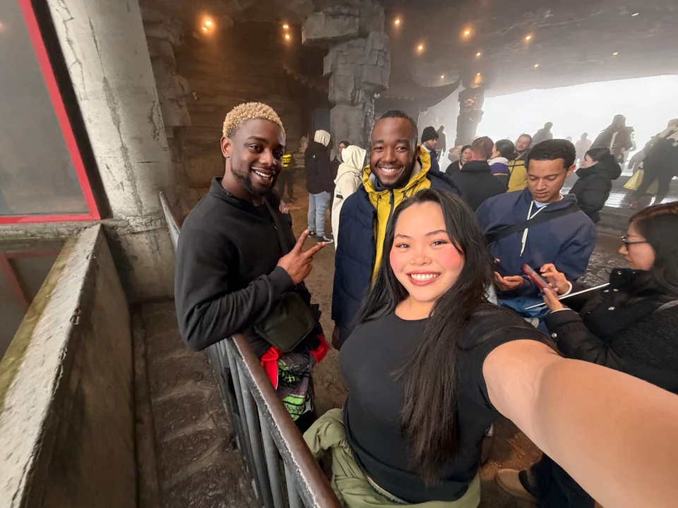Three friends taking a selfie beside a large window, with fellow travelers behind them